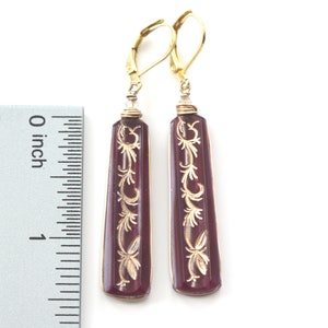 Op de afbeelding: Een paar gouden oorhangers met een paarse glazen hanger met een gegraveerd gouden bloemenmotief. De oorhangers zijn ongeveer 2,5 cm lang.