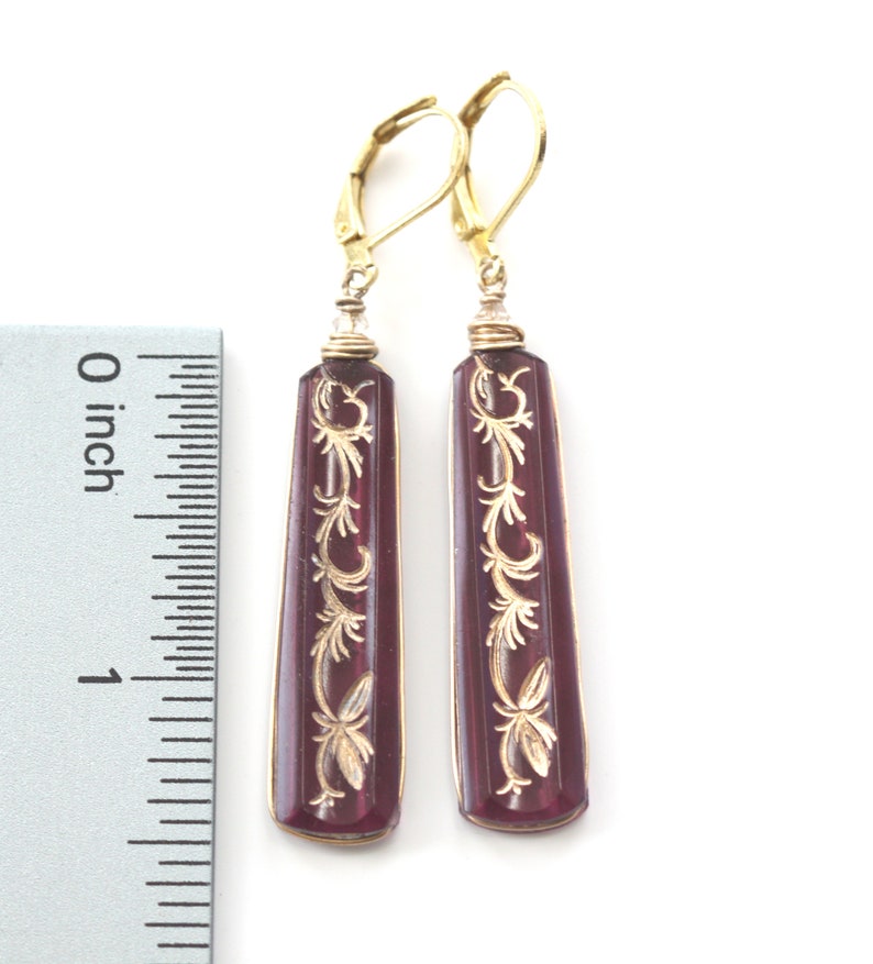 Op de afbeelding: Een paar gouden oorhangers met een paarse glazen hanger met een gegraveerd gouden bloemenmotief. De oorhangers zijn ongeveer 2,5 cm lang.