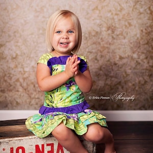 Peut inclure: Une jeune fille aux cheveux blonds est assise sur une caisse en bois avec le mot "EVEN-UP" peint dessus. Elle porte une robe à fleurs verte et violette et a un grand sourire.