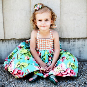 May include: A young girl wearing a blue and white floral dress with a peach and white polka dot bodice. She is sitting on a gravel surface with a white wooden wall behind her.