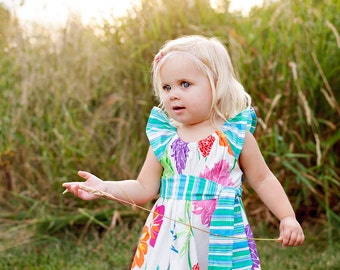 May include: A young girl with blonde hair wearing a white dress with a colorful floral pattern and a blue and green striped sash. She is standing in a field of tall grass and holding a blade of grass in her hand.