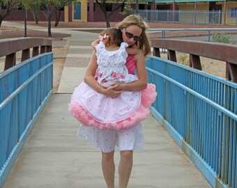May include: A woman in a pink and white dress holds a young girl in a pink and white dress with a pink tulle skirt. The woman is smiling and the girl is looking down. They are standing on a blue bridge.