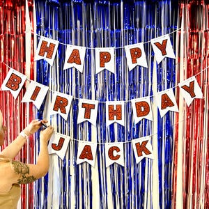 Puede incluir: Una pancarta blanca con las palabras "HAPPY BIRTHAY JACK" en letras naranjas con dise&ntilde;os de telara&ntilde;as. La pancarta est&aacute; colgada contra un tel&oacute;n de fondo de flecos met&aacute;licos rojos, blancos y azules.