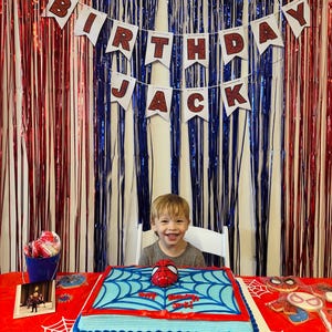 Puede incluir: Escena de fiesta de cumplea&ntilde;os con una pancarta que dice "Feliz Cumplea&ntilde;os Jack". Un ni&ntilde;o sonr&iacute;e frente a una tarta con tem&aacute;tica de Spider-Man. La tarta es azul con detalles rojos y una m&aacute;scara de Spider-Man encima. La mesa est&aacute; cubierta con un mantel rojo con un dise&ntilde;o de telara&ntilde;a.