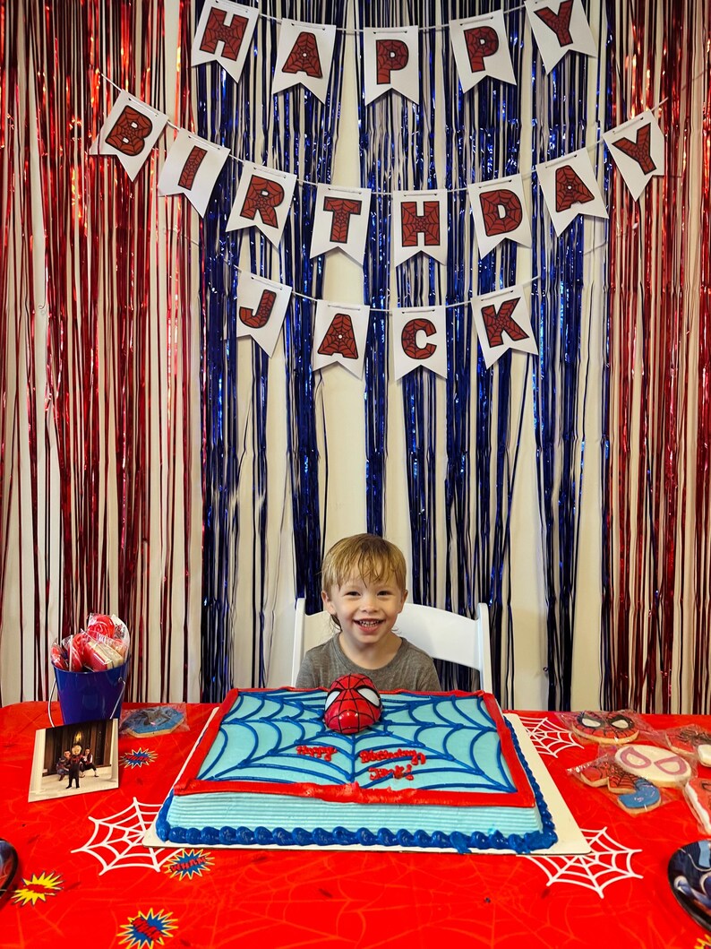Puede incluir: Escena de fiesta de cumplea&ntilde;os con una pancarta que dice "Feliz Cumplea&ntilde;os Jack". Un ni&ntilde;o sonr&iacute;e frente a una tarta con tem&aacute;tica de Spider-Man. La tarta es azul con detalles rojos y una m&aacute;scara de Spider-Man encima. La mesa est&aacute; cubierta con un mantel rojo con un dise&ntilde;o de telara&ntilde;a.