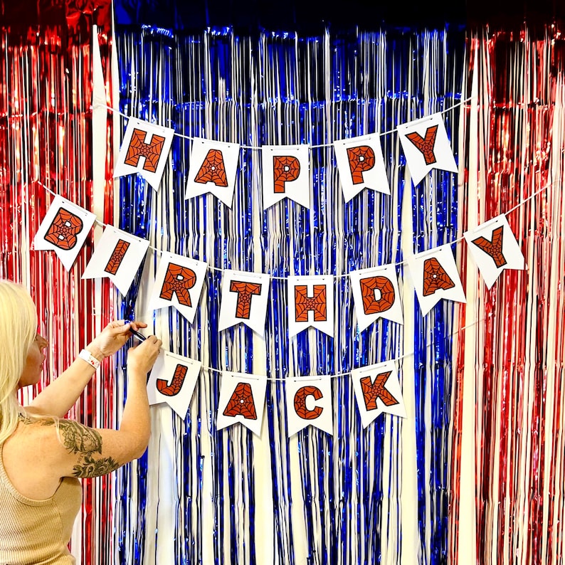 Puede incluir: Una pancarta blanca con las palabras "HAPPY BIRTHAY JACK" en letras naranjas con dise&ntilde;os de telara&ntilde;as. La pancarta est&aacute; colgada contra un tel&oacute;n de fondo de flecos met&aacute;licos rojos, blancos y azules.
