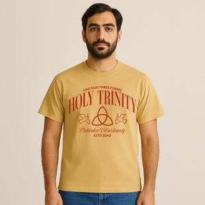 May include: Mustard yellow t-shirt featuring the red text "ONE GOD THREE FORMS HOLY TRINITY Orthodox Christianity ESTD 33AD". The design includes a trinity symbol and doves. The shirt is short-sleeved and made of a soft material.