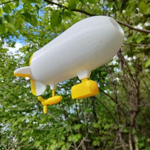 以下が含まれることがあります： 緑の葉と青い空を背景に、白いと黄色の玩具飛行船が細い糸から吊り下げられています。飛行船は黄色のフィン、プロペラ、着陸装置を備えています。おもちゃは空中に吊り下げられており、遊び心のあるアイテムまたは装飾品であることを示唆しています。