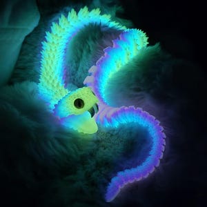 May include: A bioluminescent articulated snake toy featuring a segmented body with a vibrant gradient of glowing neon blue, green, and purple light. The snake has a textured, spiky dorsal ridge, a wide head with a dark eye, and an open mouth.