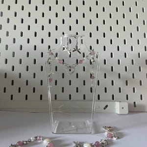 Könnte beinhalten: Ein Set aus rosafarbenem und silbernem Schmuck mit einer Halskette, Ohrringen und zwei Armbändern. Der Schmuck ist auf einer durchsichtigen Plastikpuppe ausgestellt. Der Schmuck ist mit rosafarbenen Perlen, silbernen Anhängern und einem Herzanhänger verziert.