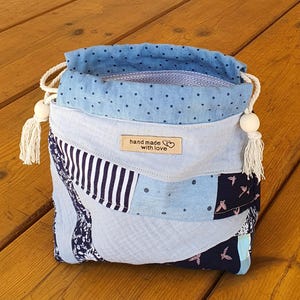 Könnte beinhalten: Ein Stoffbeutel mit Kordelzug im Patchwork-Design aus blauen, marineblauen und weißen Textilien mit Blumen-, Streifen- und Punktemustern. Ein beiges Etikett auf der Vorderseite trägt die Aufschrift "hand made with love" mit einem Herzsymbol.