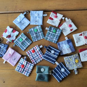 Könnte beinhalten: Eine Sammlung kleiner Stoffbeutel mit Schlüsselringen, die verschiedene Muster wie Streifen, Karos, Blumen und Erdbeerdrucke aufweisen. Jeder Beutel hat eine Klappe mit einem Kunststoff-Druckknopf und ein rundes Logo-Etikett mit der Aufschrift Lily's.
