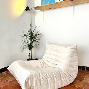 Op de afbeelding: Crèmekleurige, gewatteerde loungestoel met een laag profiel. De stoel staat in een hoek met een hoge plant in pot en een wandlamp. Boven de stoel staat een houten plank met kunst.