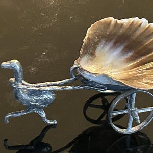 May include: A decorative miniature carriage with a seashell as the carriage bed. The carriage is pulled by a bird-like figure and has two small wheels. The seashell is beige and brown.