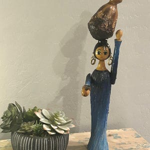 May include: A wooden figurine of a woman carrying a vessel on her head, dressed in a blue gown. The figurine has gold earrings and a painted face. A small succulent plant in a black and white striped pot is to the left.