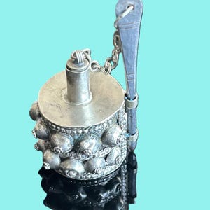 May include: A silver-toned, ornate container with a cylindrical body and a lid, connected by a chain to a small, decorative tool. The container is adorned with rounded, textured embellishments. The background is a solid teal color.