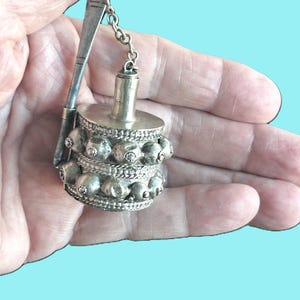 May include: A silver-toned tea caddy spoon with a decorative, round tea caddy. The caddy is adorned with multiple rows of small, round embellishments. A small chain connects the spoon to the caddy.