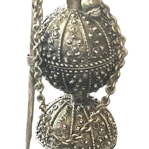 May include: A silver-toned metal container with a spherical top and a bell-shaped base, both featuring a textured surface with small raised studs. A metal chain connects the top to the base, and a long, thin metal rod is attached to the side.