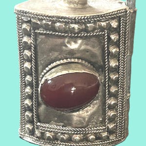May include: A silver-toned metal pendant with a rectangular body and a rounded top. The pendant features a large, oval, reddish-brown stone set in the center. The edges are adorned with intricate beaded and braided detailing.
