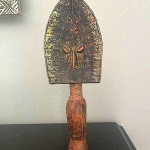 May include: A wooden sculpture with a carved face and a geometric pattern. The sculpture is painted in shades of green, brown, and black.