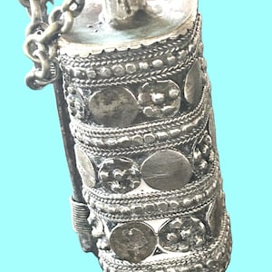 May include: A silver-toned, cylindrical pendant with intricate embossed patterns and a chain. The pendant features a series of decorative bands with circular and floral designs. The piece has a vintage aesthetic.