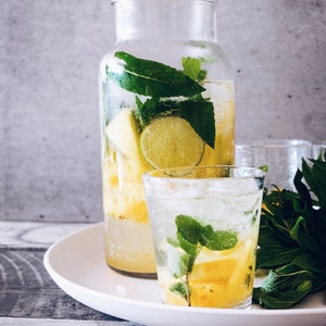 Könnte beinhalten: Ein klarer Glaskrug gefüllt mit Wasser, Ananas, Limette und Minzblättern. Ein Glas des gleichen Getränks steht auf einem weißen Teller neben dem Krug.