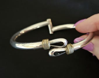 May include: A silver-toned hinged bangle bracelet featuring a polished finish and two overlapping ends. Each end is accented with a textured gold-toned band and a smooth, rounded loop design. The bracelet has a sturdy metal hinge mechanism.