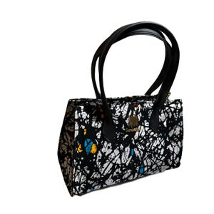 May include: A black and white patterned handbag with black handles. The bag features a unique abstract design with splashes of color. The handles are made of black leather. The bag has a gold emblem that says "Sonia R".