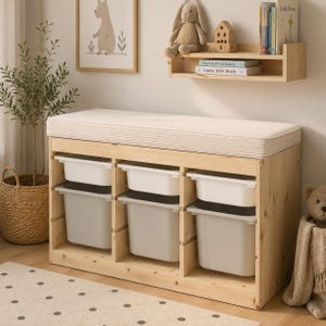 May include: A light-colored wooden storage bench featuring a thick, cream-colored fabric cushion on top. The unit includes three vertical sections, each containing one white plastic bin and one light gray plastic bin for organized storage.