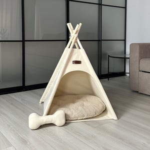 Puede incluir: Un tipi beige para mascotas con una cama de felpa y una almohada en forma de hueso. La tienda tiene un marco de madera y una cubierta de lona. La cama y la almohada son de color beige a juego. El tipi está diseñado para mascotas pequeñas.
