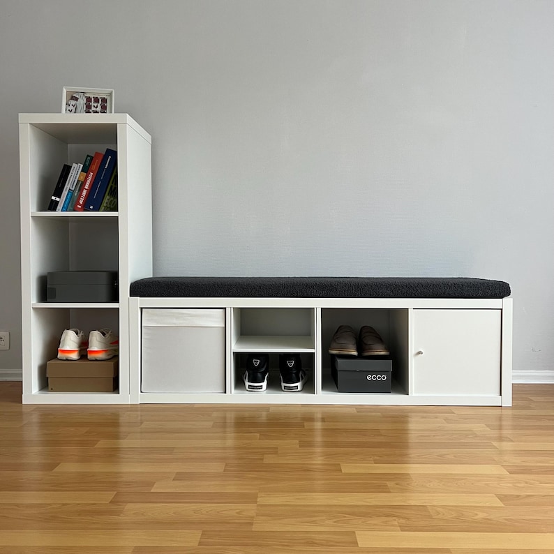 May include: White storage unit with a dark gray cushioned bench seat. The unit has open shelves, a cabinet door, and cubby spaces. Various items are stored inside, including shoes, books, and a storage bin. The unit sits on a wooden floor.