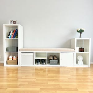 May include: A white storage unit with multiple compartments, including shelves, a drawer, and a cabinet. A beige cushion sits atop the central section. Shoes, books, and decorative items are displayed within the unit. The unit is set against a light gray wall.