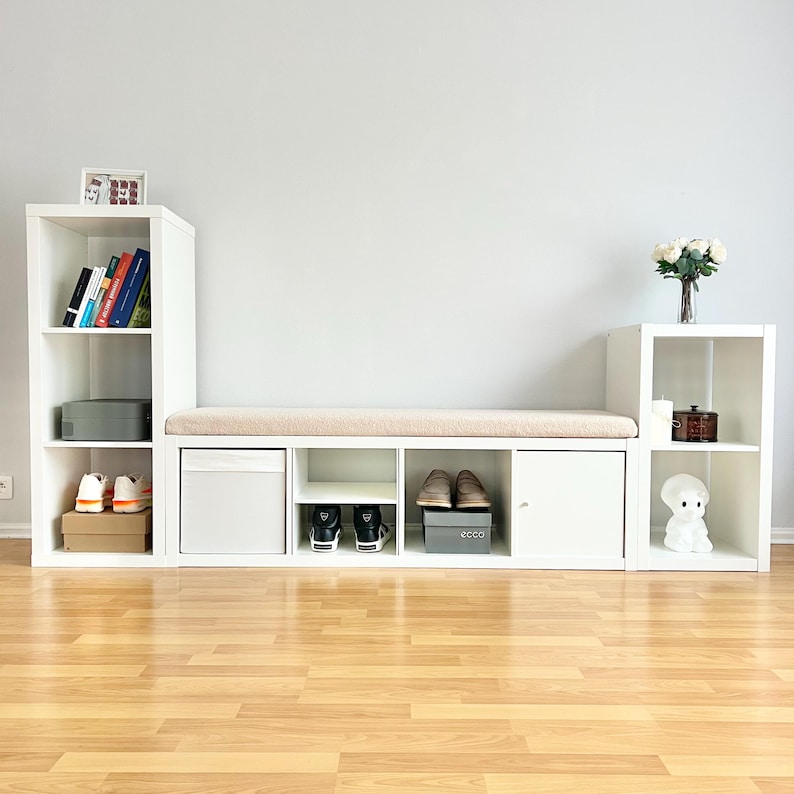 May include: A white storage unit with multiple compartments, including shelves, a drawer, and a cabinet. A beige cushion sits atop the central section. Shoes, books, and decorative items are displayed within the unit. The unit is set against a light gray wall.