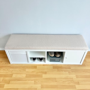 May include: A white shoe bench with a beige cushioned top. The bench has multiple storage compartments, including open shelves and a cabinet. Shoes are visible in some compartments. The bench is placed against a light gray wall.
