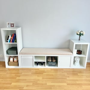 May include: White modular storage unit with a bench seat, shelves, and cabinets. The bench has a beige cushion. Shelves display books, shoes, and decorative items. The unit is against a light gray wall.