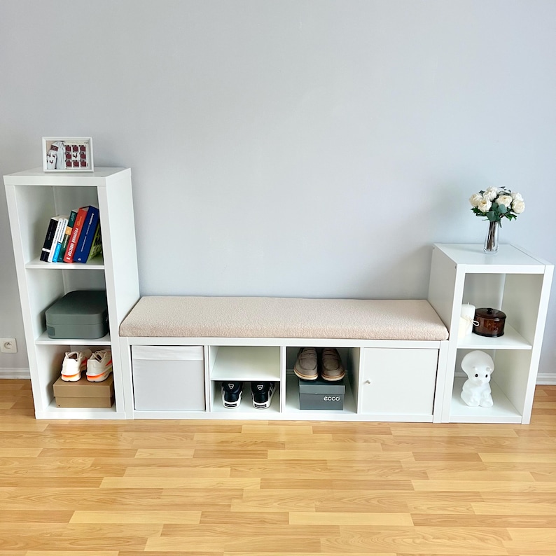 May include: White modular storage unit with a bench seat, shelves, and cabinets. The bench has a beige cushion. Shelves display books, shoes, and decorative items. The unit is against a light gray wall.