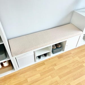 May include: A white shoe storage bench with a beige cushioned top. The bench has open shelves and a cabinet door. Shoes are visible in the open compartments and on top of the bench. The bench is against a white wall.