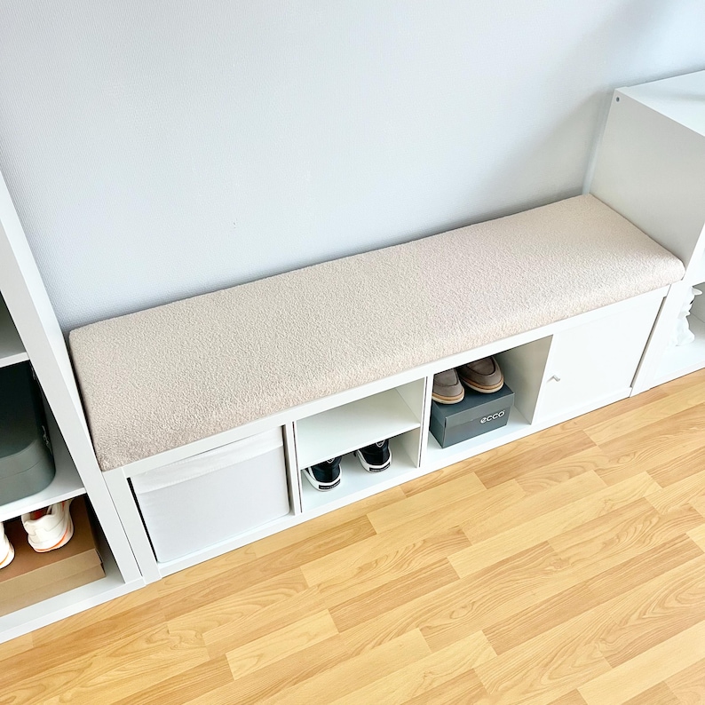 May include: A white shoe storage bench with a beige cushioned top. The bench has open shelves and a cabinet door. Shoes are visible in the open compartments and on top of the bench. The bench is against a white wall.