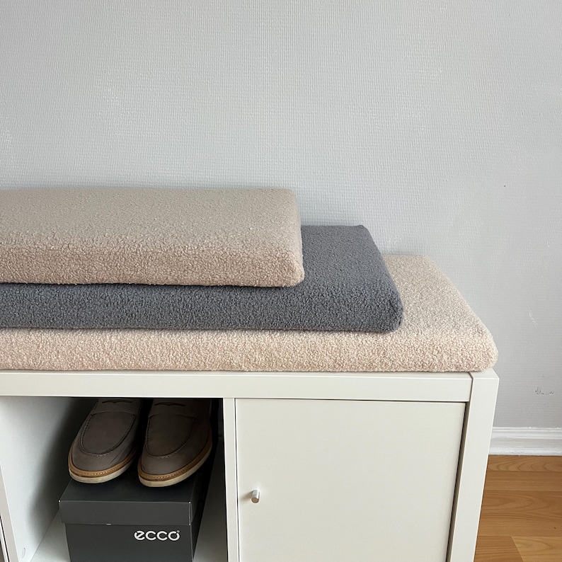 May include: Three textured, rectangular cushions in beige and gray are stacked on a white cabinet. Below, a pair of brown leather shoes sits on top of a black shoe box with the brand name "ecco" printed on it.