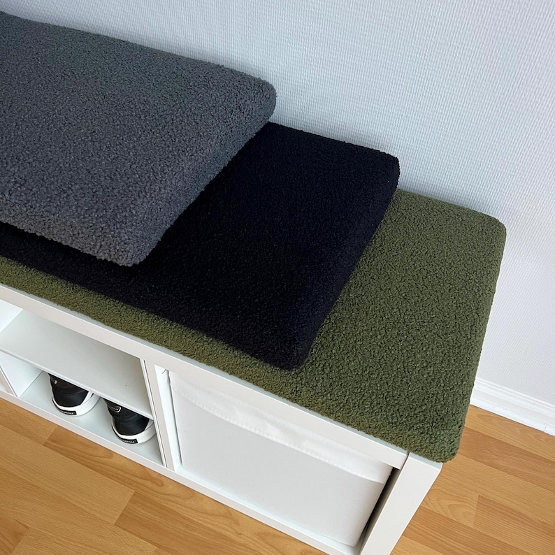 May include: Three textured bench cushions in shades of gray, black, and olive green. The cushions are stacked on a white storage bench with shoe storage. The cushions have a soft, plush appearance.