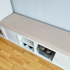 May include: A white storage bench with a beige cushioned top. The bench has open shelves and a cabinet door. Black and white sneakers and a pair of brown shoes in a box are visible in the storage compartments. The bench is placed against a light blue wall.