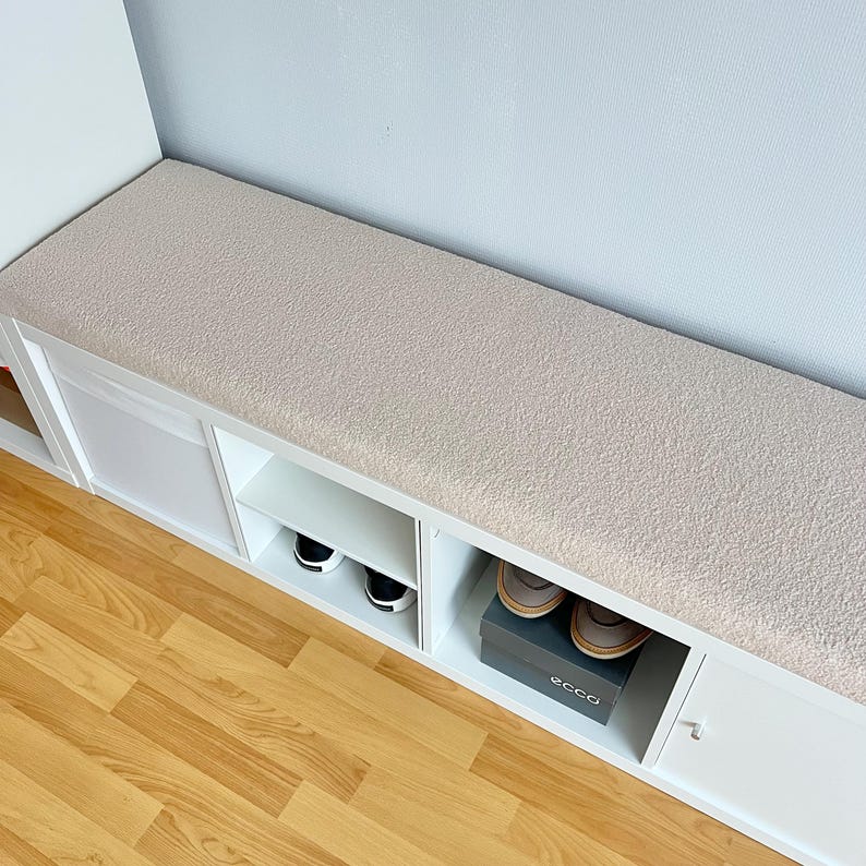 May include: A white storage bench with a beige cushioned top. The bench has open shelves and a cabinet door. Black and white sneakers and a pair of brown shoes in a box are visible in the storage compartments. The bench is placed against a light blue wall.