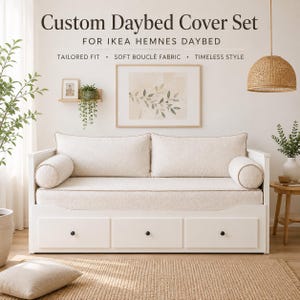 Peut inclure: Un lit de repos blanc avec une housse sur mesure en tissu bouclé crème, comprenant un long coussin d'assise, deux coussins de dossier rectangulaires et deux traversins cylindriques. Le cadre possède trois tiroirs de rangement à boutons noirs.