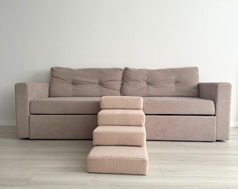 May include: A beige pet step with four steps is positioned in front of a matching beige sofa. The steps are made of a soft, textured material. The sofa has a button-tufted back and a low profile.