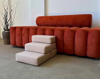May include: A modern, burnt orange sofa with a cylindrical bolster pillow and a set of three beige steps. The sofa has a ribbed design. A large vase with branches is on the left.