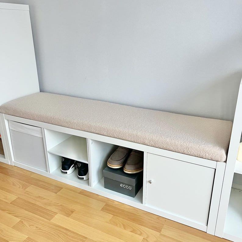 Puede incluir: Un banco de almacenamiento blanco con una parte superior acolchada beige. El banco tiene compartimentos abiertos y una puerta de armario. Los zapatos son visibles dentro de los compartimentos. El banco est&aacute; dise&ntilde;ado para la organizaci&oacute;n y los asientos.