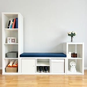 May include: White modular storage unit with a blue cushion bench, shelves, and storage compartments. Includes books, shoes, and decorative items. The unit is against a white wall and has a wooden floor.