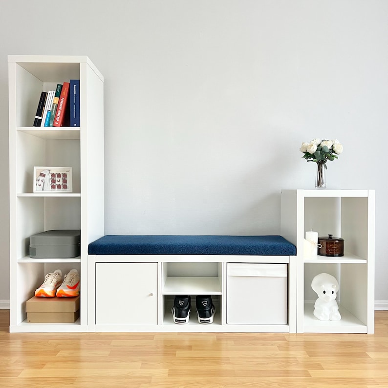 May include: White modular storage unit with a blue cushion bench, shelves, and storage compartments. Includes books, shoes, and decorative items. The unit is against a white wall and has a wooden floor.