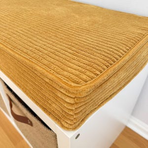 May include: A rectangular mustard yellow corduroy cushion with a thick, ribbed texture and piped edges, positioned on top of a white storage unit that features a square cubby containing a beige woven basket with a brown leather handle.