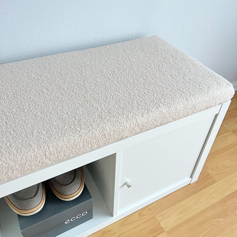 May include: A white shoe bench with a plush, cream-colored cushioned top. The bench has a storage compartment with a door and an open shelf. A pair of brown shoes sits on a shoe box on the shelf.